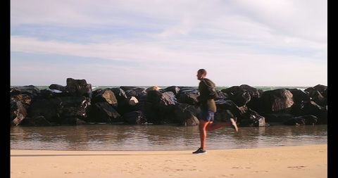 African American Man Jogging on Sandy Beach by Rock Barrier