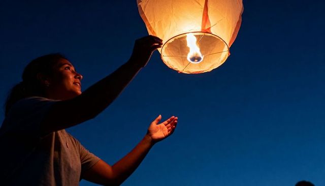 Releasing glowing paper lantern into twilight sky, woman celebrating warm flame