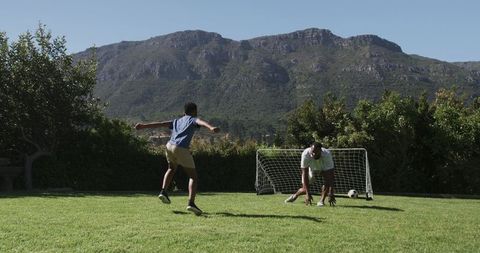 Backyard soccer practice: boy taking shot at adult goalkeeper against mountain backdrop