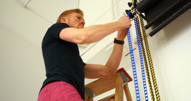 Young man is seen adjusting equipment while standing on a ladder in an industrial setting. He is wearing casual attire and appears focused on his task. Useful for illustrating concepts involving work, diligence, and technical skills. Ideal for industry-related articles, safety guidelines, and educational materials.