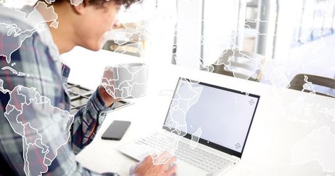 Young man working on laptop with coffee and world map overlay in sunlit coworking space