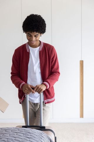 Young man standing in bright bedroom holding suitcase handle wearing red bomber jacket