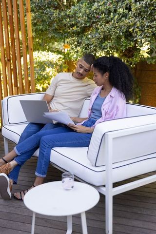 Diverse couple reviewing documents and laptop on white outdoor sofa, patio teamwork