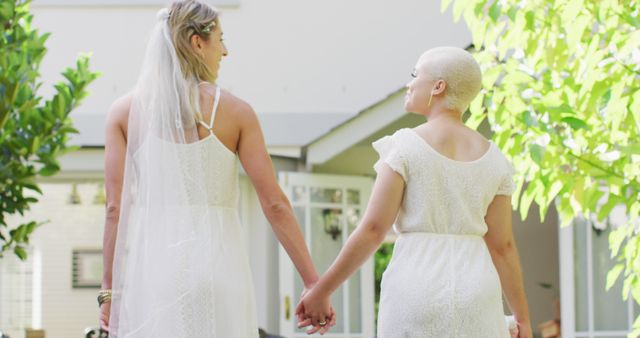 Same-sex couple wearing wedding dresses holding hands outdoors amid lush green surroundings. Perfect for themes on LGBTQ relationships, love, weddings, and unity. Can be used for wedding articles, LGBTQ advocacy, and relationship counseling materials.