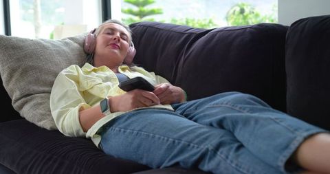 Mature woman relaxing with wireless headphones on dark sofa holding remote, cozy home