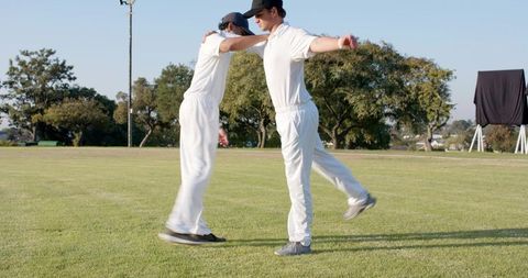 Cricket teammates celebrating by embracing and jumping on local grass field in white kit