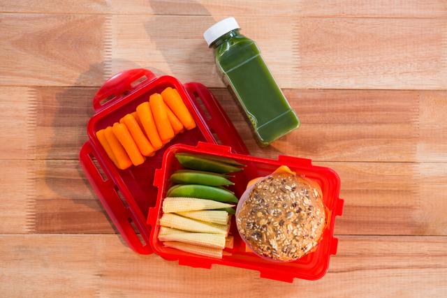 Healthy lunch box containing a whole grain sandwich, sliced cucumbers, baby carrots, corn, and a bottle of green juice on a wooden table. Ideal for promoting balanced diet, meal prep, and healthy eating habits. Suitable for use in articles, blogs, and advertisements related to nutrition, school lunches, and fitness.