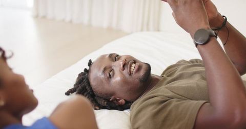 Smiling black man reclining on bed using smartphone while partner watches, lifestyle