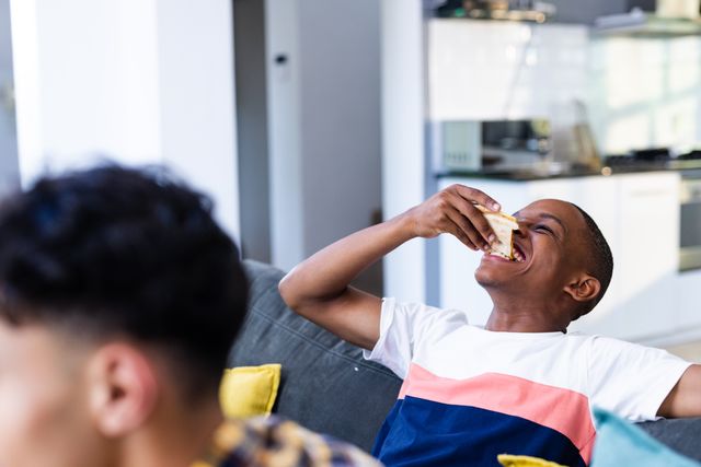 Happy african american teenage boy eating pizza slice and sitting on sofa at home. Hanging out with friends and spending quality time together concept.