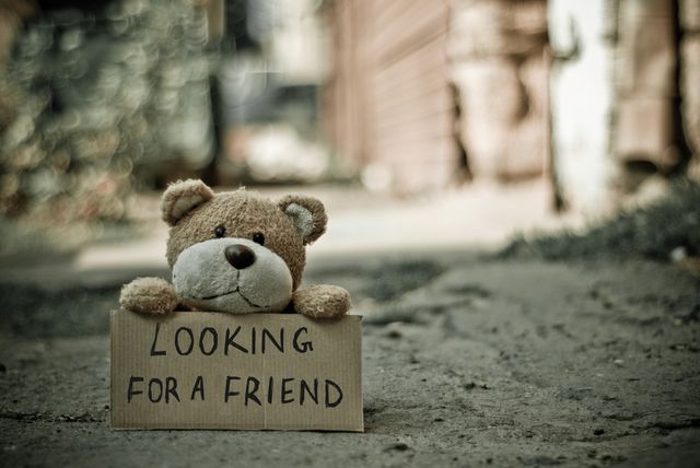 Image of lonely teddy bear holding cardboard sign 'Looking for a Friend' in urban alley. Useful for concepts like loneliness, friendship, abandonment, childhood nostalgia. Suitable for social campaigns, advertisements illustrating companionship and mental health. Great for blog posts about loneliness, adoption, and emotional appeal.
