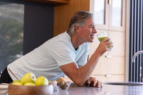 Mature man leaning on modern kitchen island drinking green smoothie for healthy lifestyle