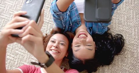 Two young female friends are lying on the floor, having fun while taking a selfie together. Their joyful expressions exhibit friendship and the enjoyment of technology. This can be used for themes involving friendship, technology use, youth interactions, and diverse moments of happiness. Ideal for social media, technology promotions, or friendship articles.