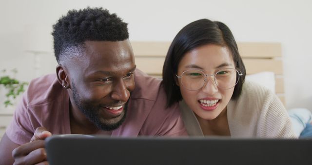 Cheerful diverse couple watching a movie together on a laptop, smiling happily. Suited for use in advertisements or articles about modern relationships, home entertainment, technology, multicultural interactions, or mutual hobbies. Perfect for marketing campaigns promoting streaming services or inclusive lifestyle.