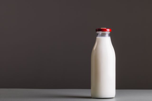 This image features a glass milk bottle with a red cap against a gray background. The minimalist composition and ample copy space make it ideal for use in advertisements, health and nutrition articles, dairy product promotions, and food-related blogs. The clean and simple design emphasizes freshness and purity.