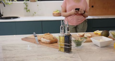 Woman checking phone while preparing sandwich on marble countertop in modern teal kitchen