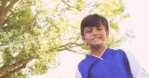 Smiling boy wearing blue soccer jersey standing under sunlit tree with string lights