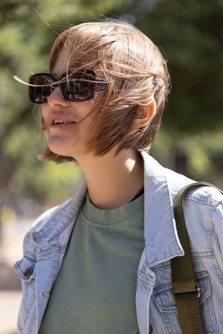 Young woman in sunlit park wearing denim jacket, tortoise sunglasses, backpack strap