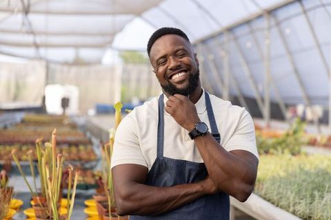 Smiling african american nursery owner standing in greenhouse wearing apron and smartwatch