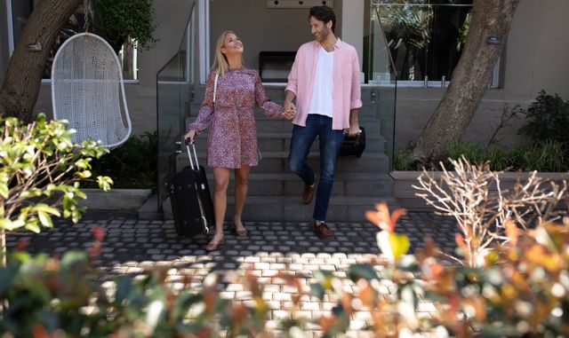 Happy Caucasian couple walking down front steps of hotel, holding hands and smiling, carrying luggage on a sunny day. Ideal for travel advertisements, vacation packages, romantic getaways, and hotel promotions.