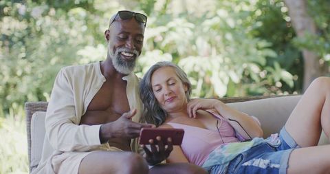 Senior couple relaxing on outdoor wicker sofa sharing smartphone and enjoying sunshine