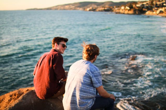 Two young men sitting on an ocean cliff, enjoying the scenic view and each other's company at sunset. This can be used for themes of friendship, travel, relaxation, and adventure. Suitable for marketing materials promoting outdoor activities, travel destinations, or leisure apparel.