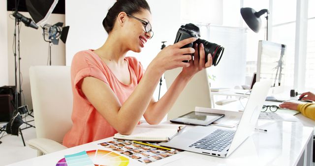 Female photographer reviewing pictures on DSLR camera while sitting in a modern office. She appears happy and engaged, surrounded by a laptop, tablet, and color palette, suggesting a creative and professional work environment. Perfect for concepts of modern photography, creative industries, professional photography, and workspace setups.