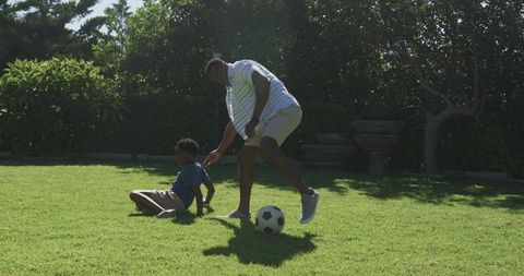 African father playing soccer with son on sunny backyard lawn, family bonding