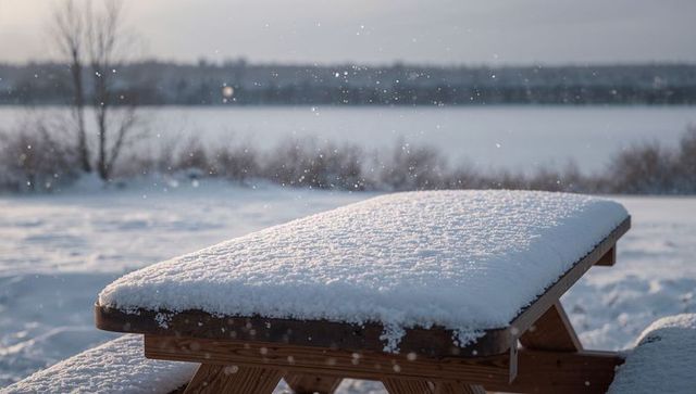 Snow-covered picnic table overlooking frozen lake, falling snowflakes, winter calm