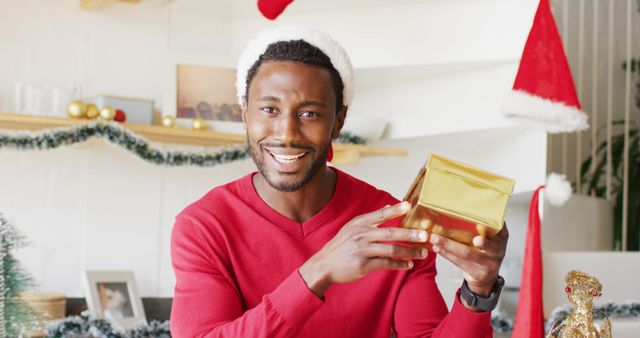 Cheerful man enjoys Christmas, showing a golden gift box while wearing Santa hat and red sweater. Perfect for holiday advertisements, festive greeting cards, blog posts about Christmas joy, and social media promotions highlighting holiday spirit and cheerful giving.