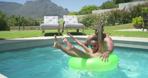 African american father and son playing on neon green inflatable tube in backyard pool with mountain
