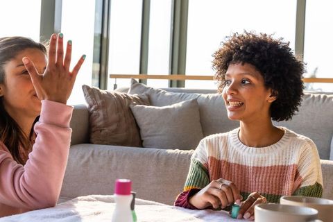 Friends painting nails at sunlit living room table smiling sharing cozy manicure moment