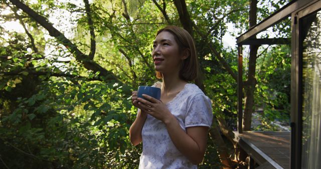 Woman holding a blue mug and enjoying a hot beverage in a lush forest environment with sunlight filtering through the trees. Ideal for use in promotions about relaxation, nature retreats, peaceful living, morning routines, and outdoor activities.
