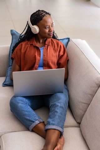 African american woman working remotely on laptop listening with headphones on beige sofa
