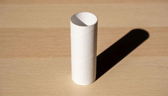 Standing cardboard tube casting long sharp shadow on light wooden tabletop minimalist