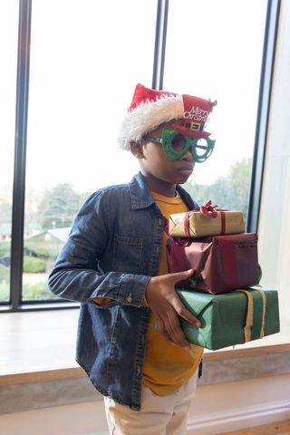 Boy wearing santa hat and green novelty glasses holding stack of wrapped christmas gifts