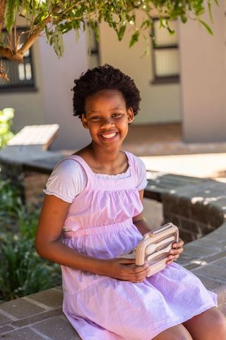 Smiling african american girl in pink dress sitting on brick wall holding beige case