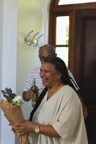 Laughing couple celebrating new home with bouquet and wine in sunlit foyer entryway