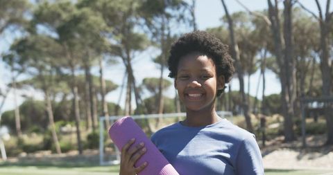 Young woman holding purple yoga mat in park smiling outdoor fitness and wellness
