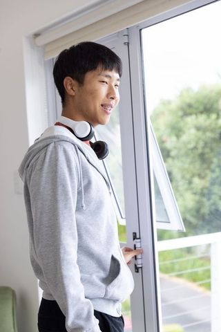 Young asian man standing by balcony door wearing hoodie and headphones, looking out at greenery
