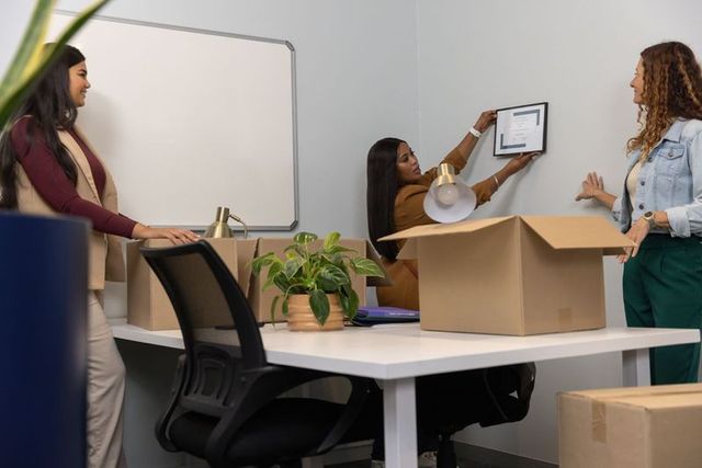 Diverse female coworkers unpacking boxes and hanging certificate in new office corner