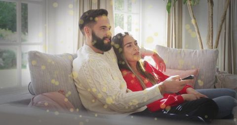 Couple cuddling on sunlit living room sofa wearing cozy sweaters holding TV remote relaxing