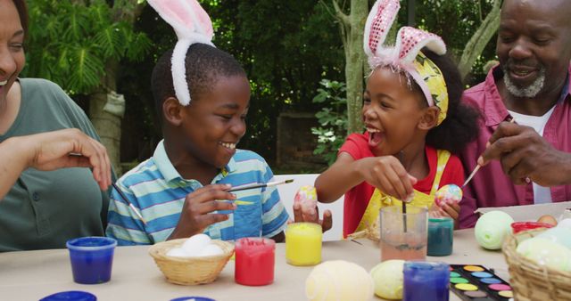 Family wearing bunny ears is painting Easter eggs with paints. Children are enjoying crafts at a table outdoors, smiling and laughing together. This happy scene is perfect for promoting family-oriented products, holiday campaigns, and creative activities for kids, emphasizing fun and togetherness.
