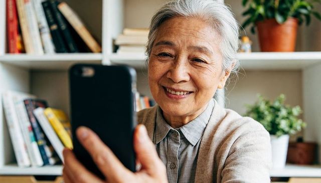 Senior asian woman holding smartphone smiling on video call at home with books and plants