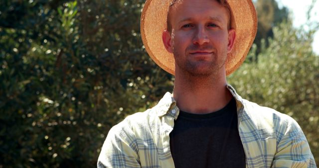 This image shows a confident man wearing a straw hat and a plaid shirt standing in a sunlit garden with trees in the background. The natural light suggests a pleasant and warm day, making it a suitable choice for themes related to outdoor activities, gardening, rural lifestyle, and self-assurance. It could be used in lifestyle blogs, websites focusing on nature or outdoor activities, and promotional content for garden-related products or services.