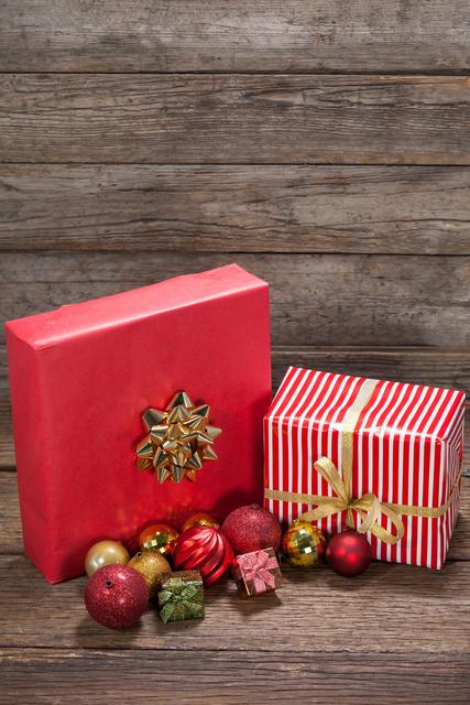 Christmas gifts wrapped in red paper with golden ribbons and bows, accompanied by festive ornaments, are placed on a rustic wooden table. Ideal for holiday-themed promotions, greeting cards, and festive advertisements.