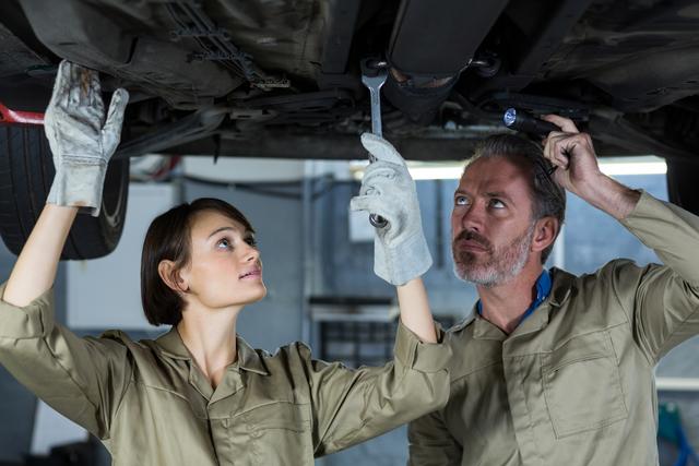 Mechanics examining car undercarriage in repair shop, using tools for maintenance and inspection. Ideal for content related to automotive repair, teamwork in workshops, professional mechanics, and car maintenance services.
