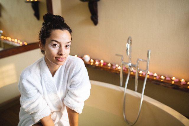 Biracial woman with vitiligo wearing a white bathrobe, sitting by a bathtub in a spa setting. Ideal for use in wellness, self-care, and beauty promotions, as well as articles on skincare and body positivity.