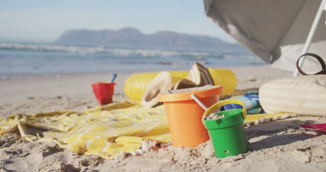 Colorful sand buckets positioned on a sandy beach with an umbrella beside them. A yellow blanket, a straw hat, and mountains in the distance add to the scenic beauty. This visual could be perfect for travel brochures, vacation promotion materials, children's toy ads, or articles about beach outings.