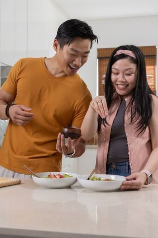 Couple preparing fresh green salads together in bright modern kitchen, seasoning and smiling
