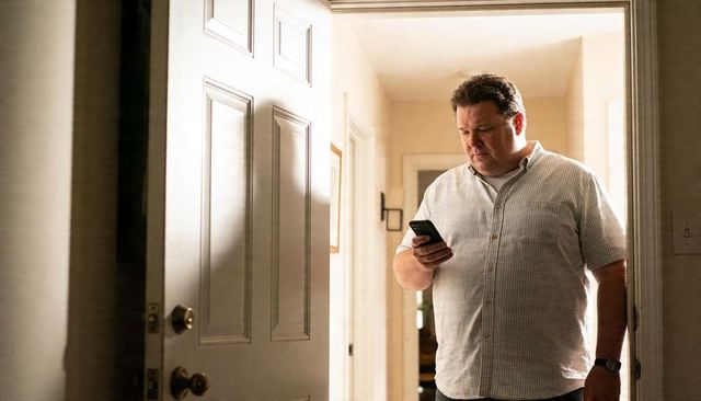 Man checking phone in home entryway by front door wearing checked shirt and watch